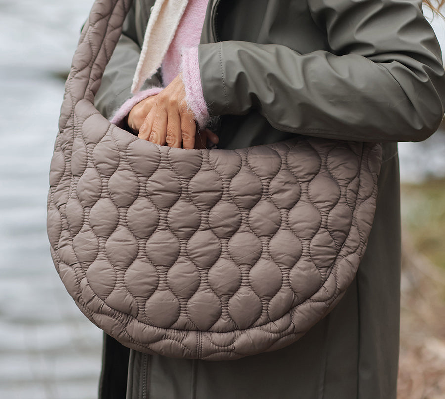 Model carrying the POM Polly beige quilted half moon bag over one arm outdoors by a lake, styled with a khaki rain jacket and pink fluffy scarf, showing the generous size and bubble quilt detail.