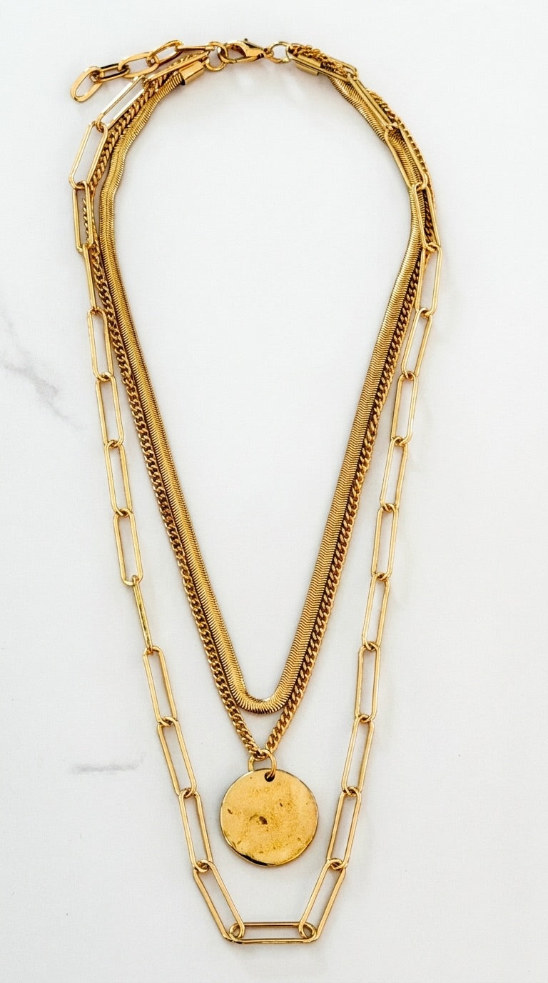 Envy gold triple layer necklace with herringbone snake chain, curb chain, paperclip chain and hammered coin pendant on marble background