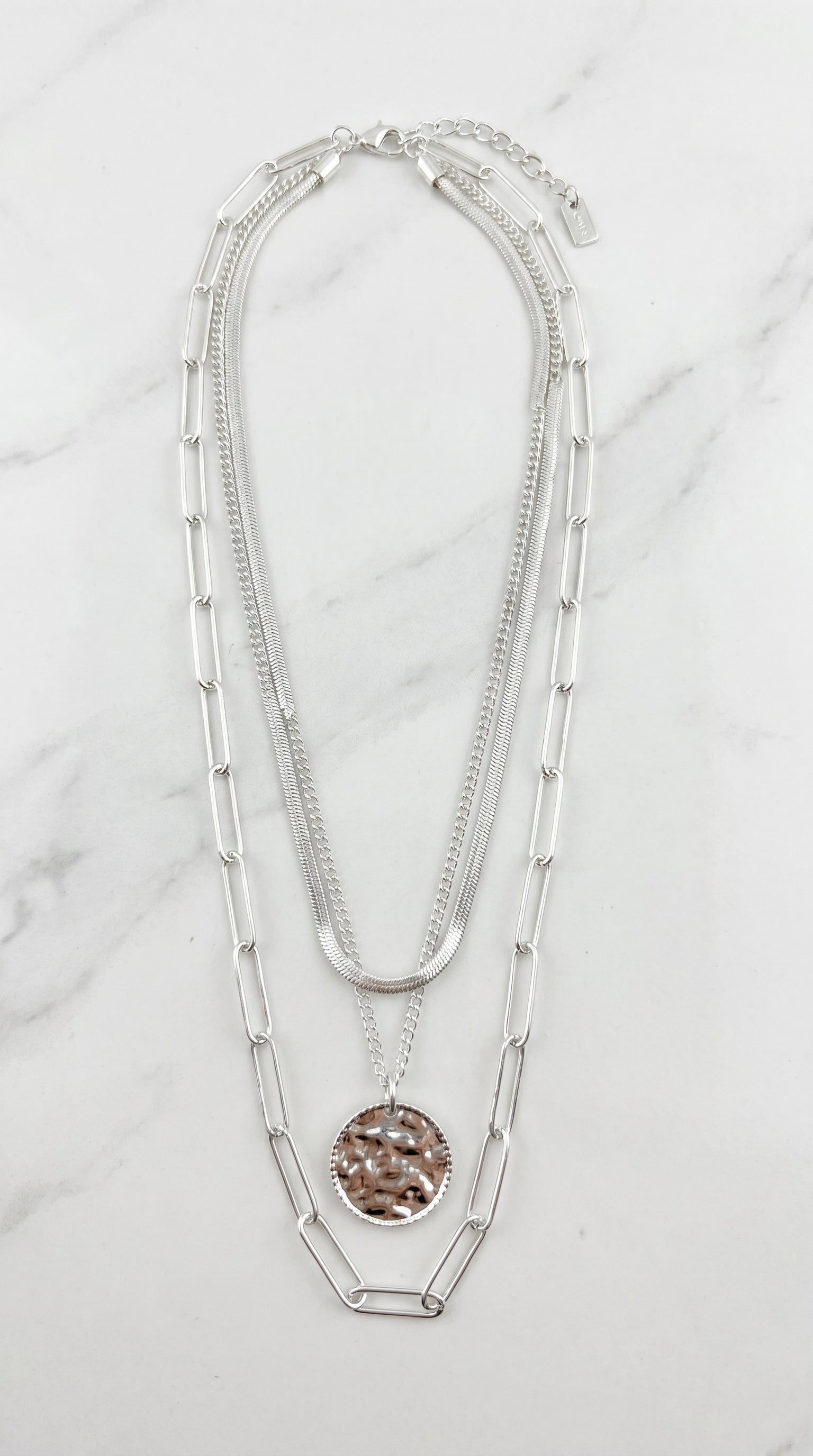 Envy silver triple layer necklace with herringbone snake chain, curb chain, paperclip chain and hammered coin pendant on marble background