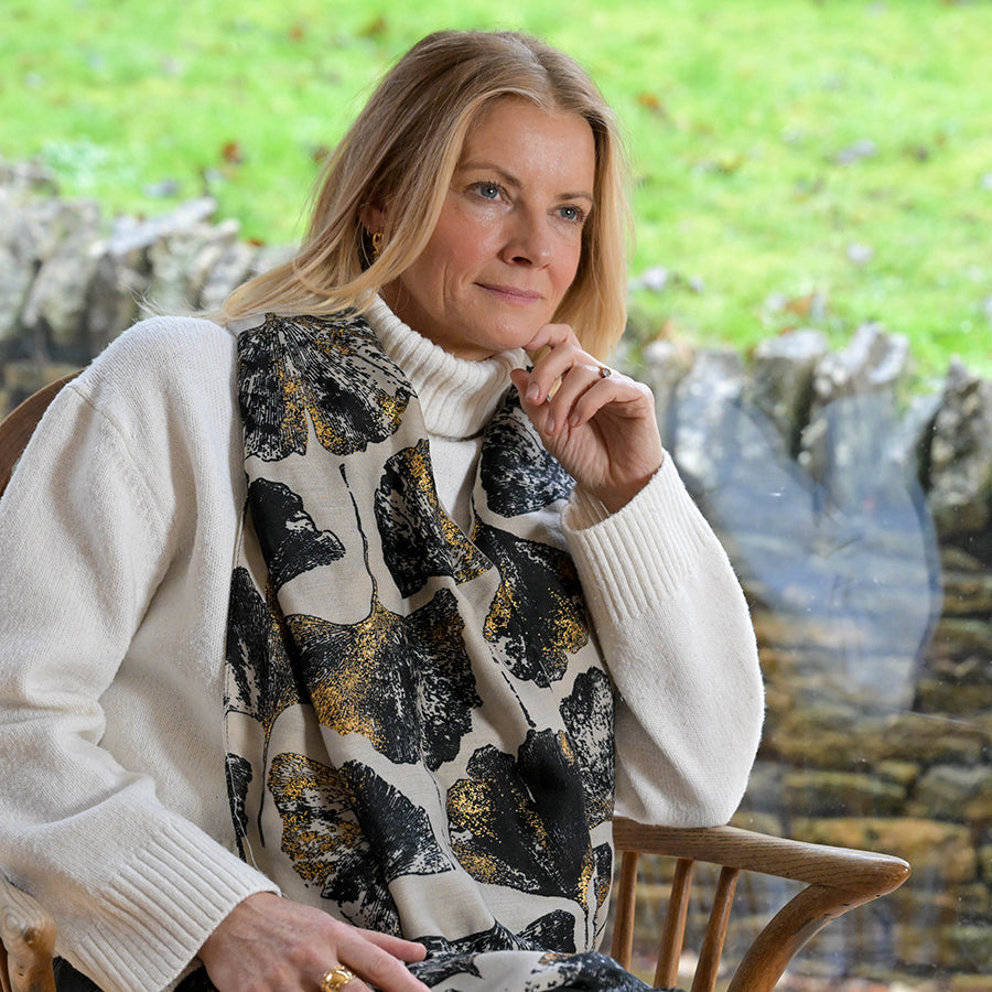 Smiling blonde model seated outdoors in a garden setting wearing the POM Greta natural mix ginkgo and metallic gold print scarf draped loosely around the neck, styled with a cream chunky knit jumper and gold hoop earrings, with a stone wall and green lawn behind.
