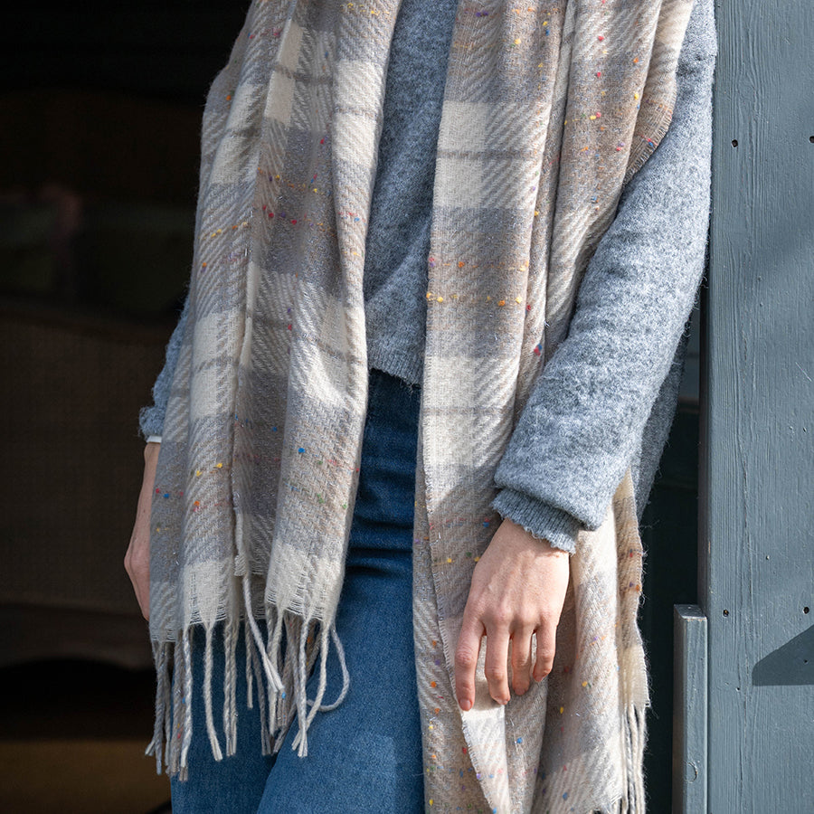 Close-up of model wearing the POM Olivia taupe mix tartan weave scarf draped open over a blue-grey knit jumper and jeans, showing the oversized taupe and cream tartan weave with multicolour confetti fleck and twisted fringe ends.