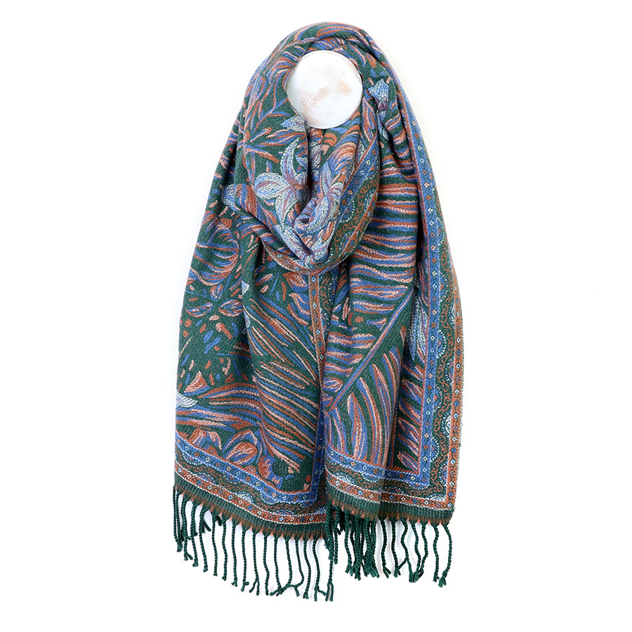 POM Celia green-blue and rust reversible jacquard scarf looped on a mannequin stand against a white background, showing the deep teal green side with large botanical floral and leaf jacquard motifs in rust, pale blue and cream, with twisted green tassel fringe ends.