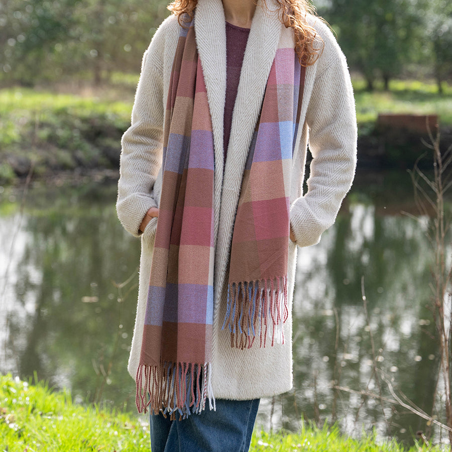 Model standing outdoors by a lake wearing the POM Esme blush and blue mix check scarf draped loosely over the shoulders, styled with a cream herringbone coat and jeans, with green trees and water in the background.