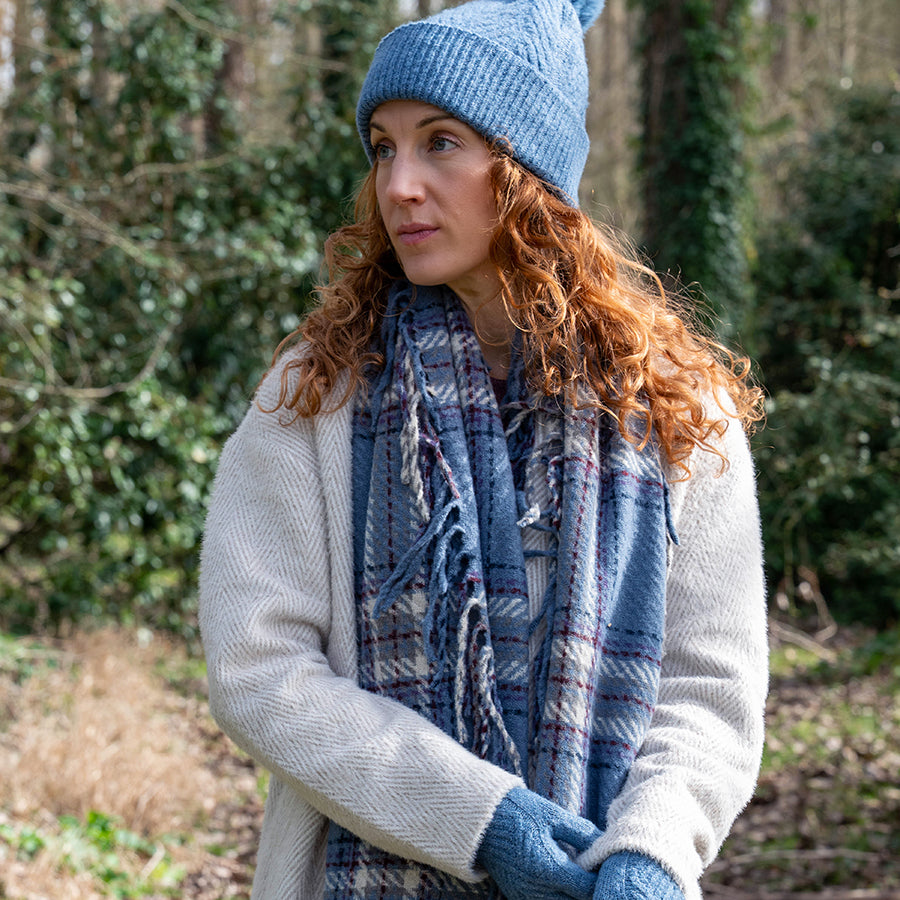 Model with curly auburn hair outdoors in a woodland setting wearing the POM Darcie dusky blue mix fringe edge check scarf wrapped around the neck, styled with a matching blue knit beanie, blue gloves and a cream herringbone coat.