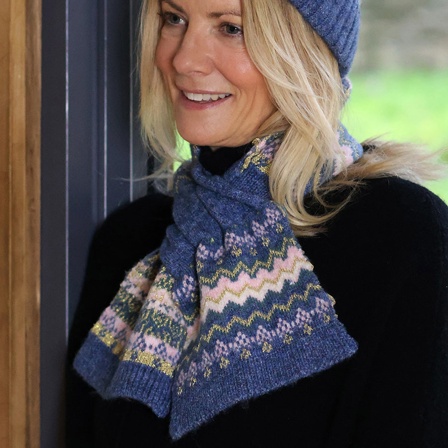 Smiling blonde model leaning in a doorway wearing the POM Sofia denim, pink and gold Fair Isle pull-through scarf threaded through at the neck, styled with the matching Sofia denim Fair Isle bobble hat and a black jumper, with a green garden behind.