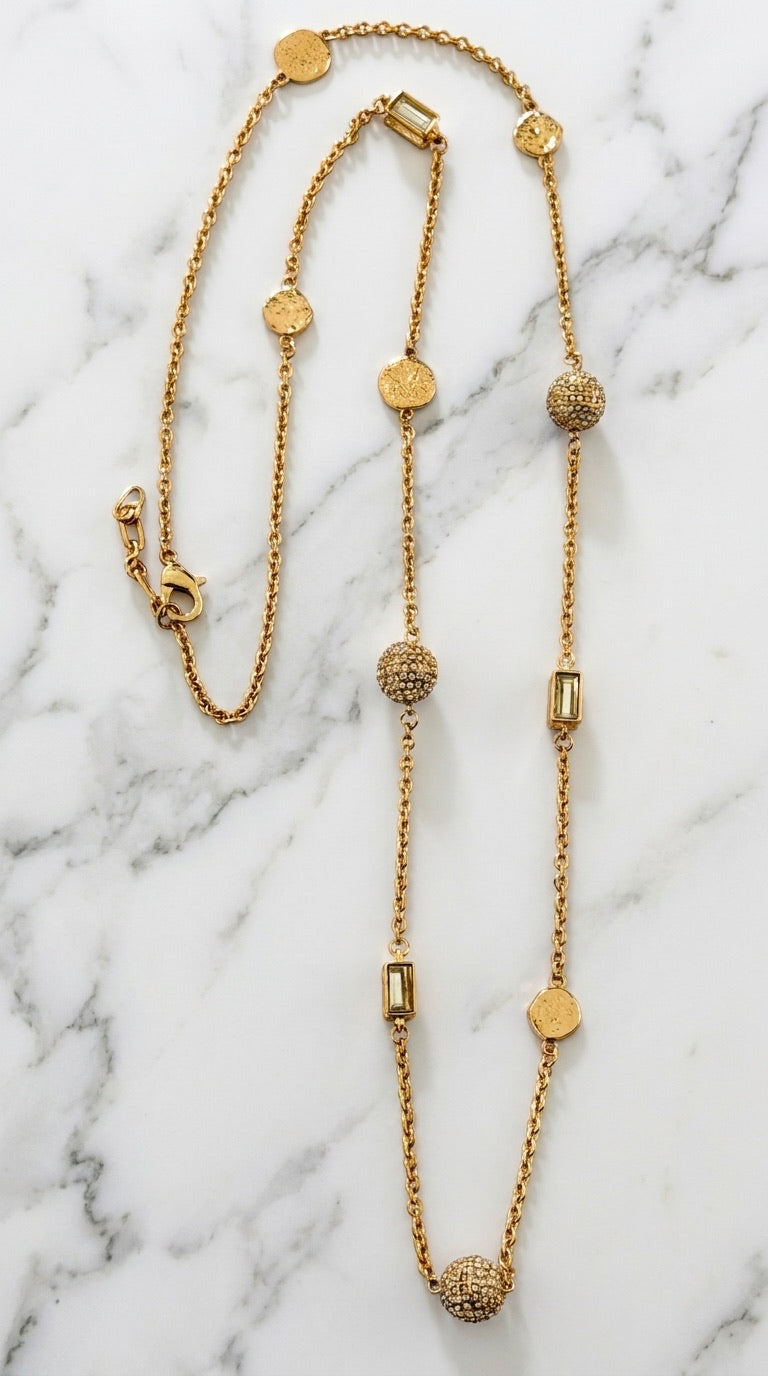 Envy long gold rolo chain necklace with pavé crystal ball, baguette, and engraved disc stations on marble background