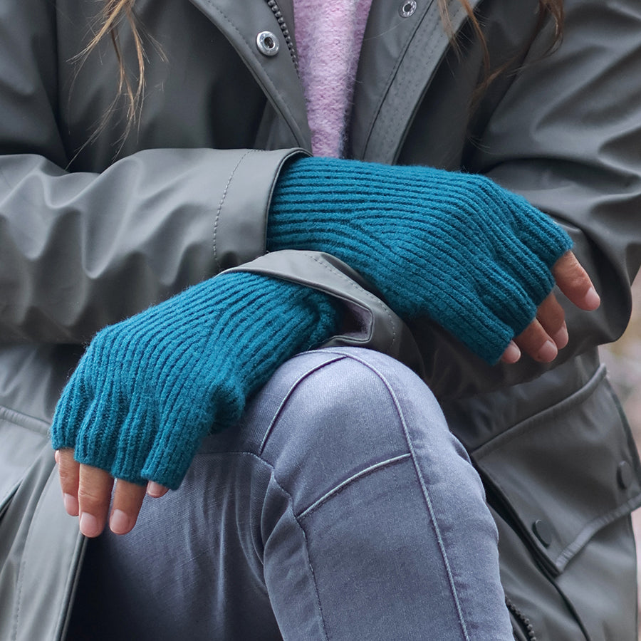 Model wearing POM Yasmin teal ribbed fingerless gloves resting on knees, styled with a grey leather jacket and lilac jumper outdoors, showing the long ribbed wrist coverage.