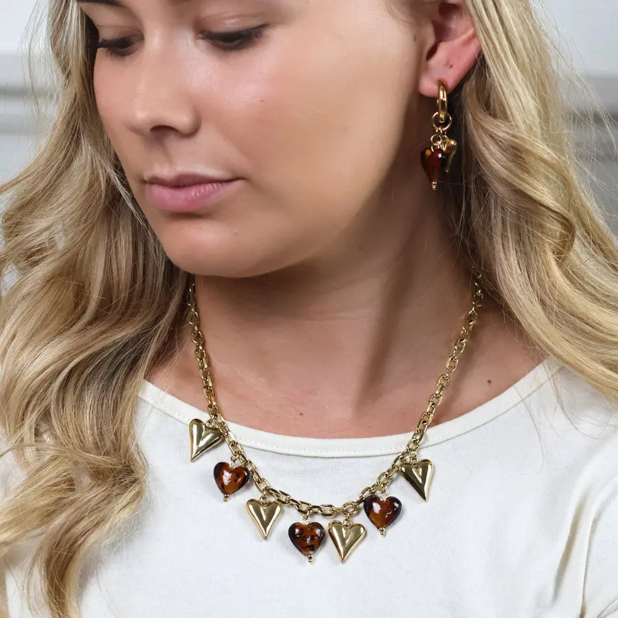 Model wearing POM Treasure gold and brown heart charm necklace with matching tortoiseshell heart drop earrings, styled with a white top.
