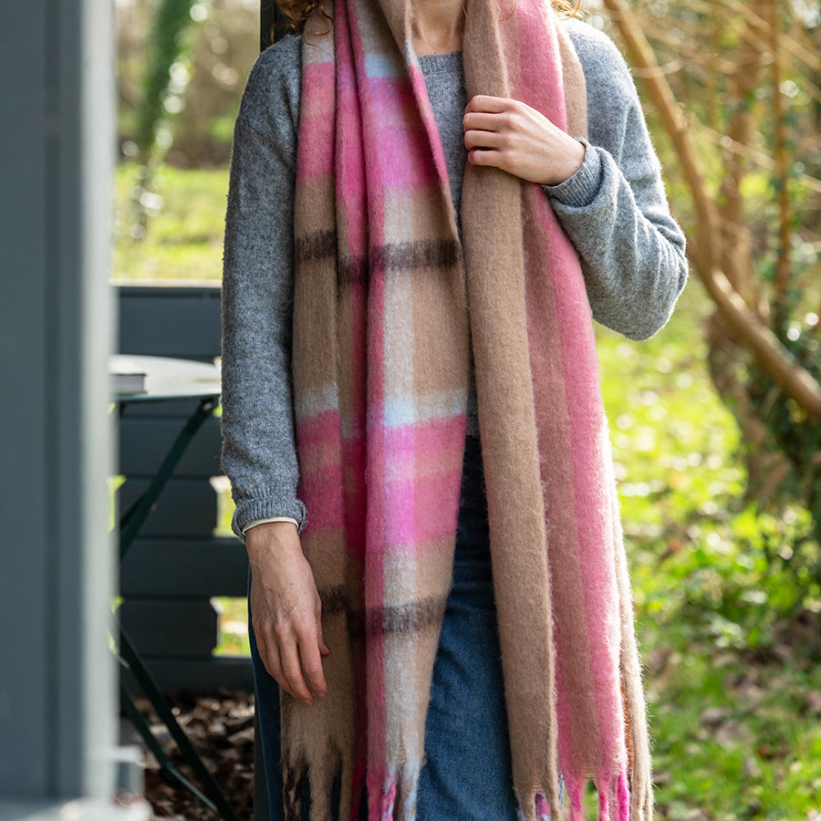 Model wearing the POM Molly taupe and pink mix check scarf draped loosely over both shoulders, styled with a grey knit jumper and jeans in an outdoor garden setting.