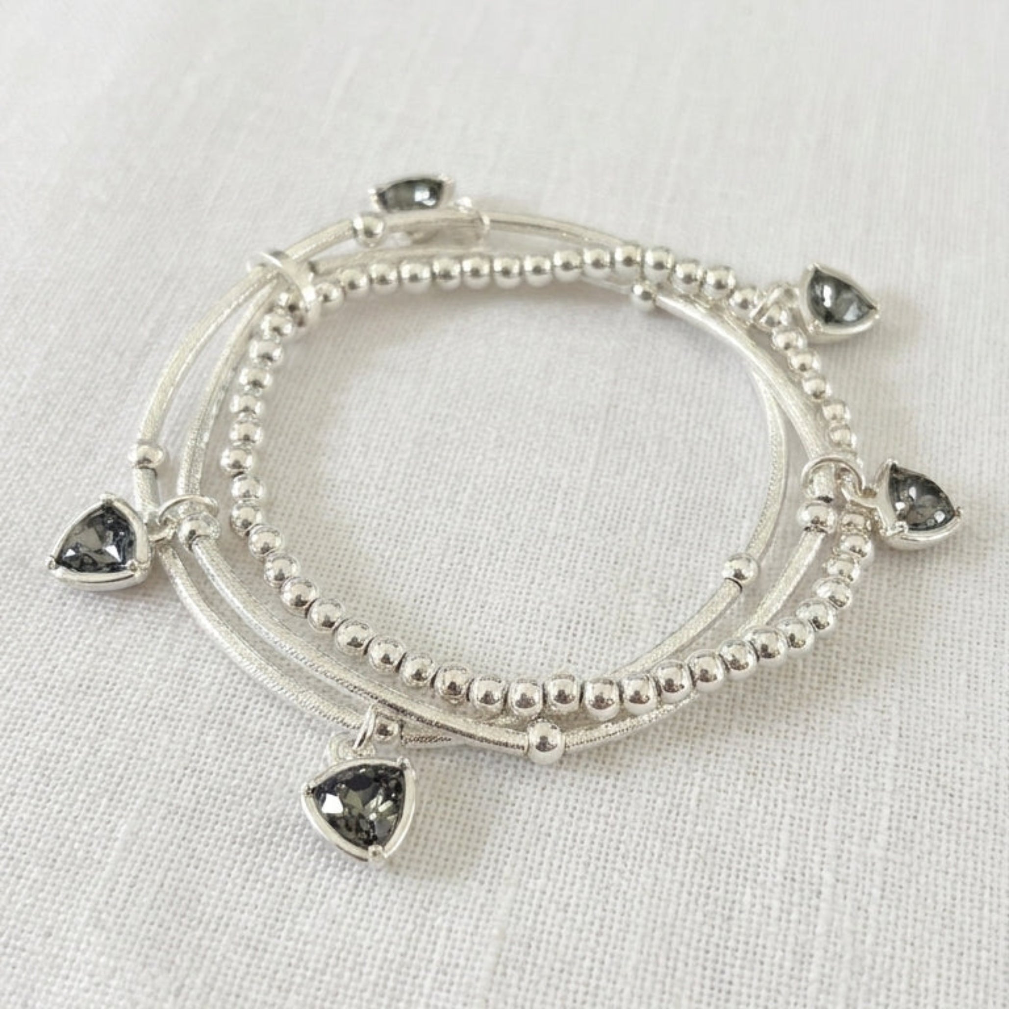 Envy silver triple wrap bracelet set with ball bead strands and smoky grey trillion-cut crystal drop charms on cream linen background