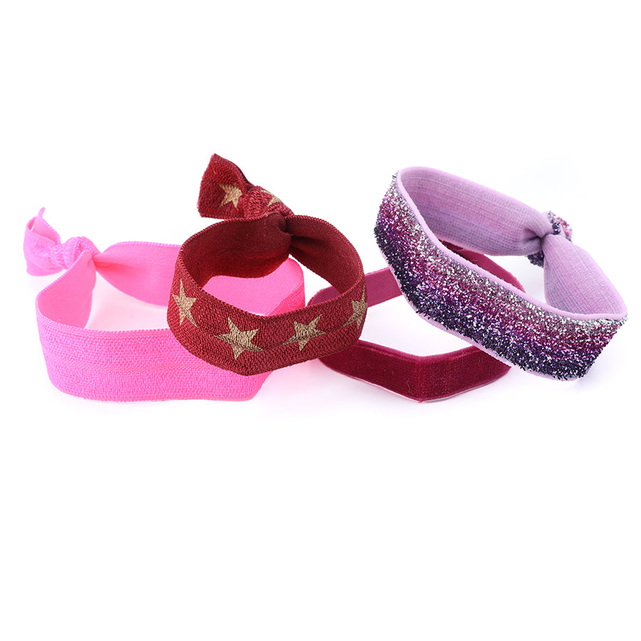 Three POM fold-over elastic hair ties worn as wristbands — hot pink, red and gold star print, and purple glitter ombré — styled together on a white surface.