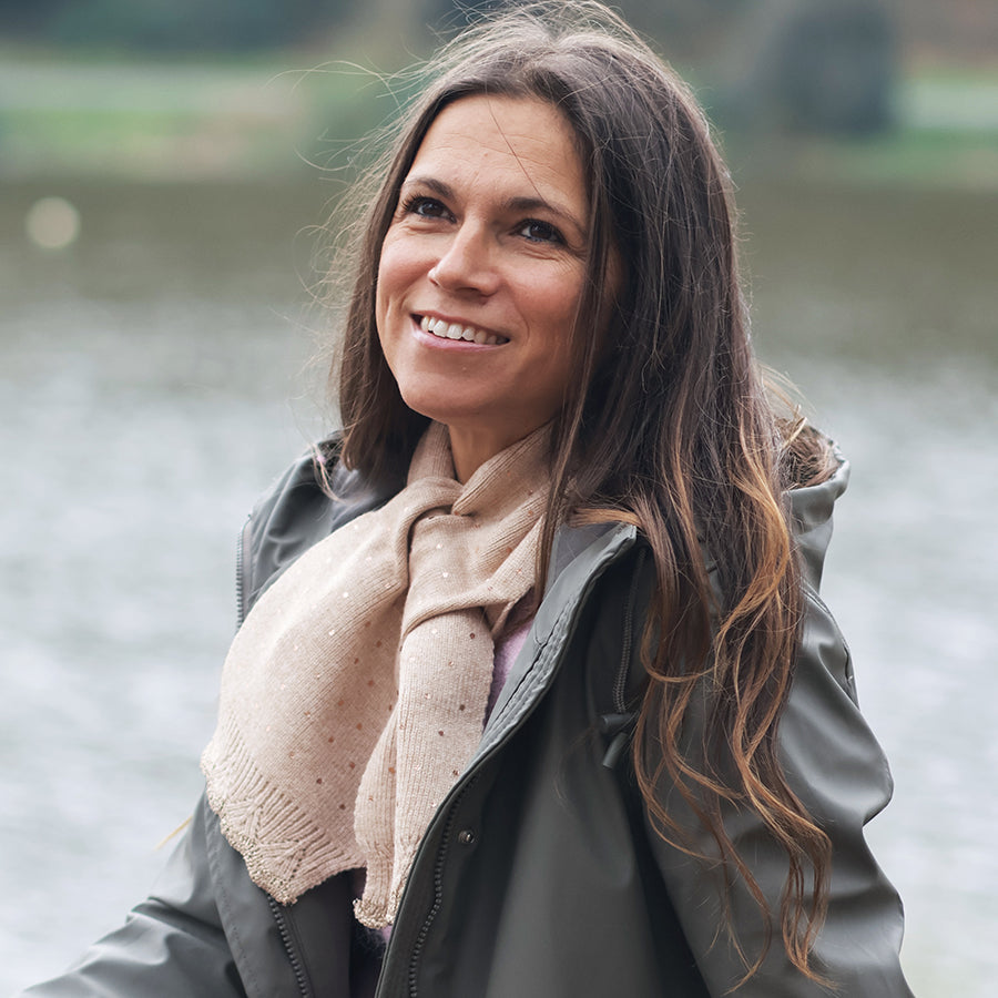 Smiling model looking upward outdoors by a lake wearing the POM Yasmin champagne beige metallic spot pull-through scarf loosely at the neck, styled with a dark grey waxed jacket and long wavy hair.