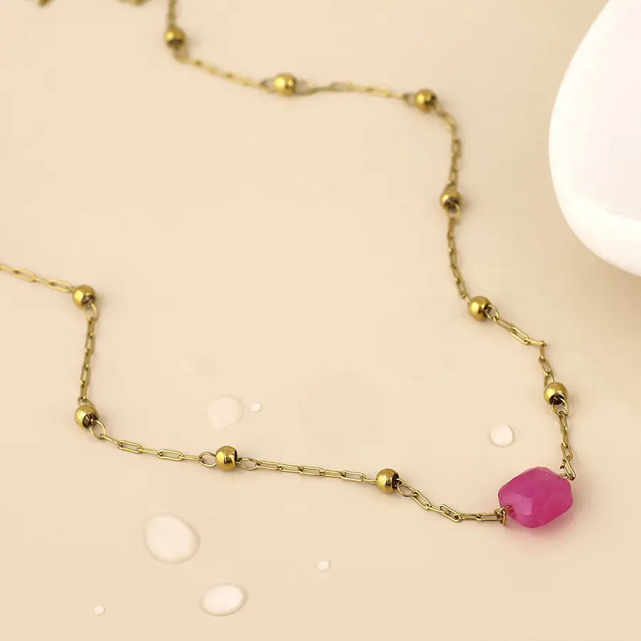A delicate gold chain necklace with small metallic beads and a faceted pink gemstone pendant, displayed on a light beige surface with water droplets.