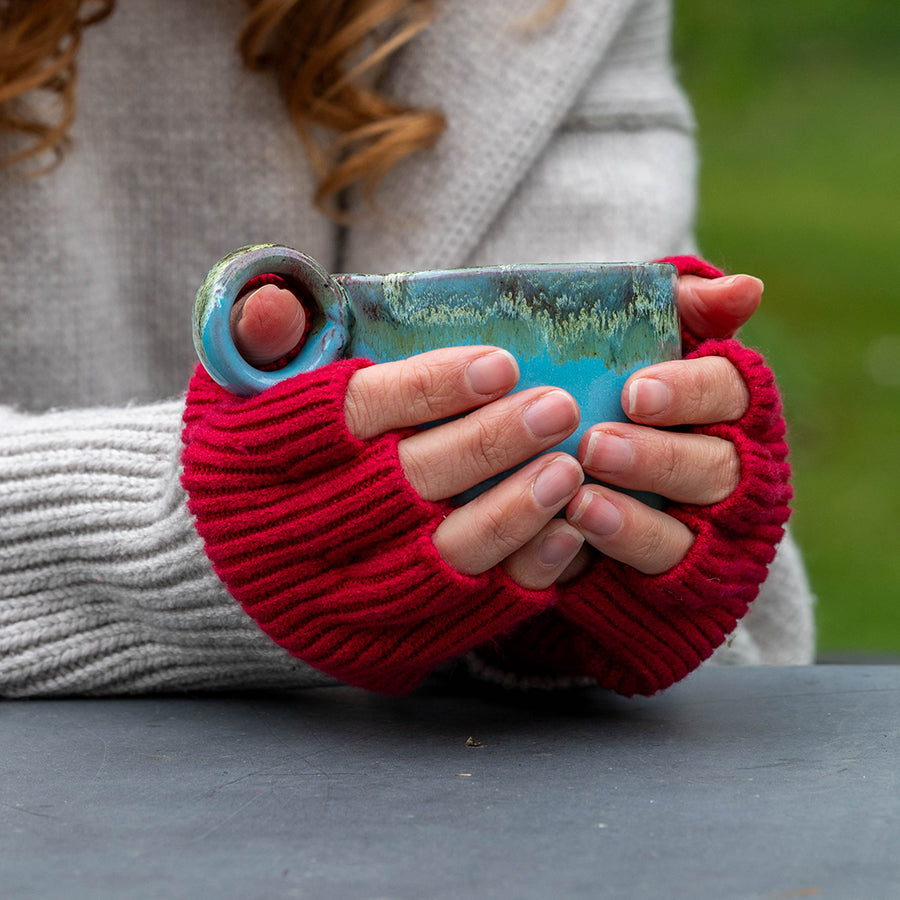 Close-up of a model's hands wearing the POM rose red fine rib knit fingerless gloves outdoors, cradling a teal ceramic mug, styled with a cream chunky knit jumper and grey coat, with a green garden background.