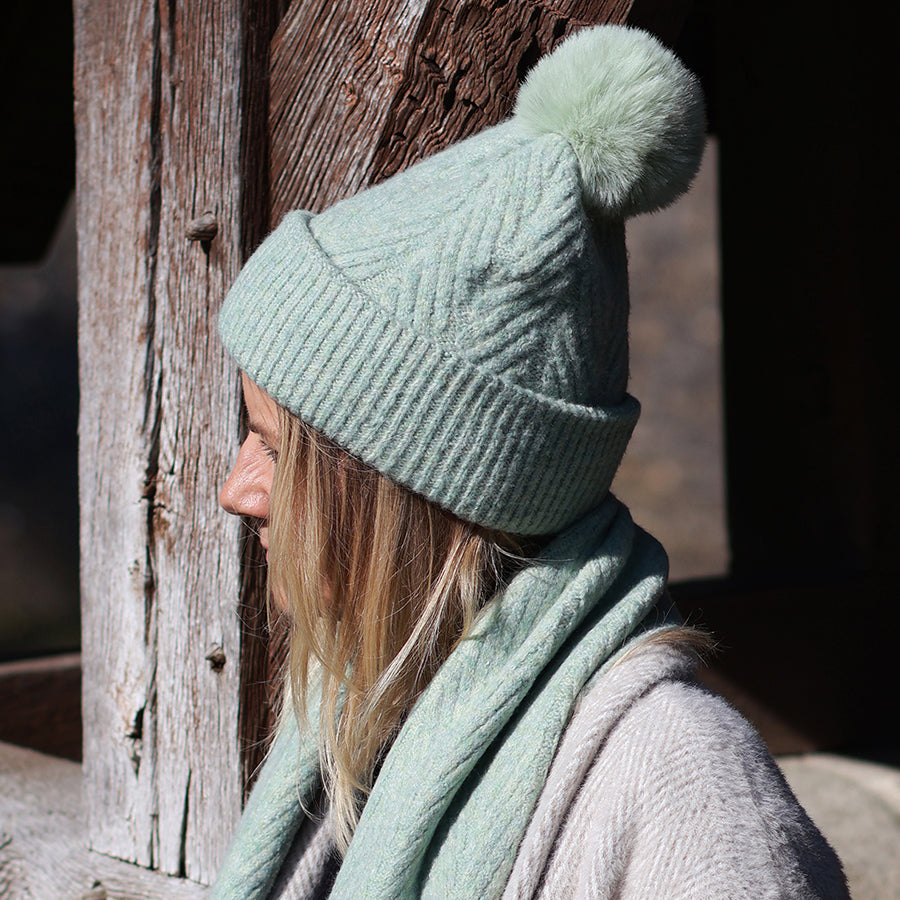 Side profile of model wearing the POM Charlotte mint herringbone knit bobble hat, styled with a matching mint scarf and a cream coat, leaning against a weathered wooden post in bright winter light.