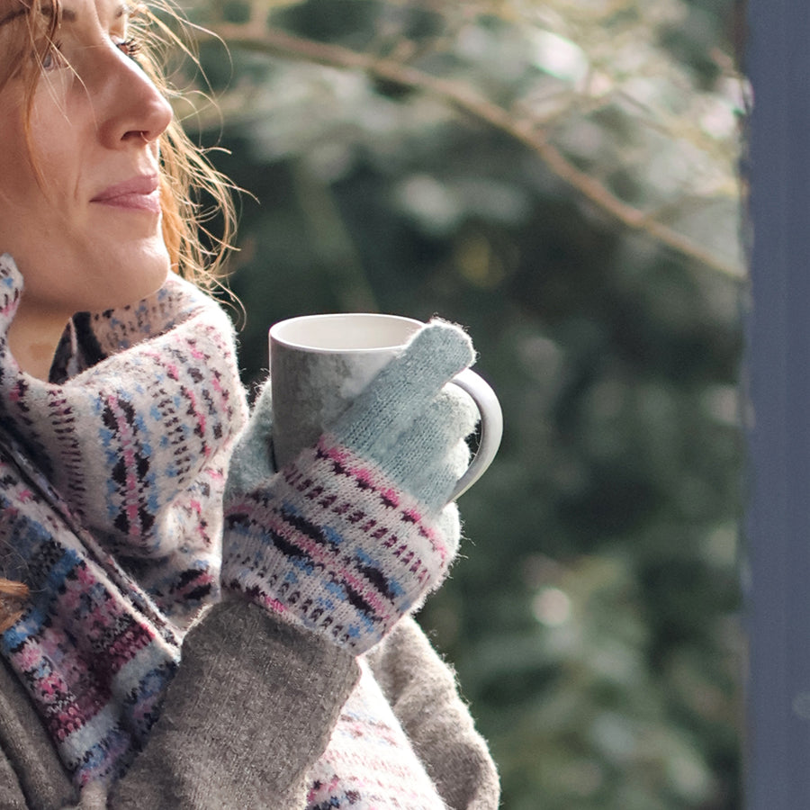 Model holding a grey ceramic mug with both hands wearing POM Iris mint fair isle knit gloves outdoors in winter sunlight, styled with a matching Iris fair isle scarf and a grey wool coat.