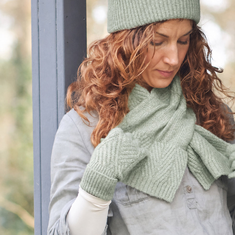 Model looking down leaning against a dark post wearing POM Charlotte light sage cable knit gloves, styled with a matching sage cable knit scarf and sage ribbed beanie, over a grey denim jacket outdoors.