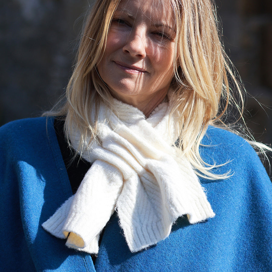 Model looking down with eyes closed wearing the POM Charlotte cream herringbone pull-through scarf, styled with a cobalt blue wool coat outdoors in bright winter sunlight against a stone wall.