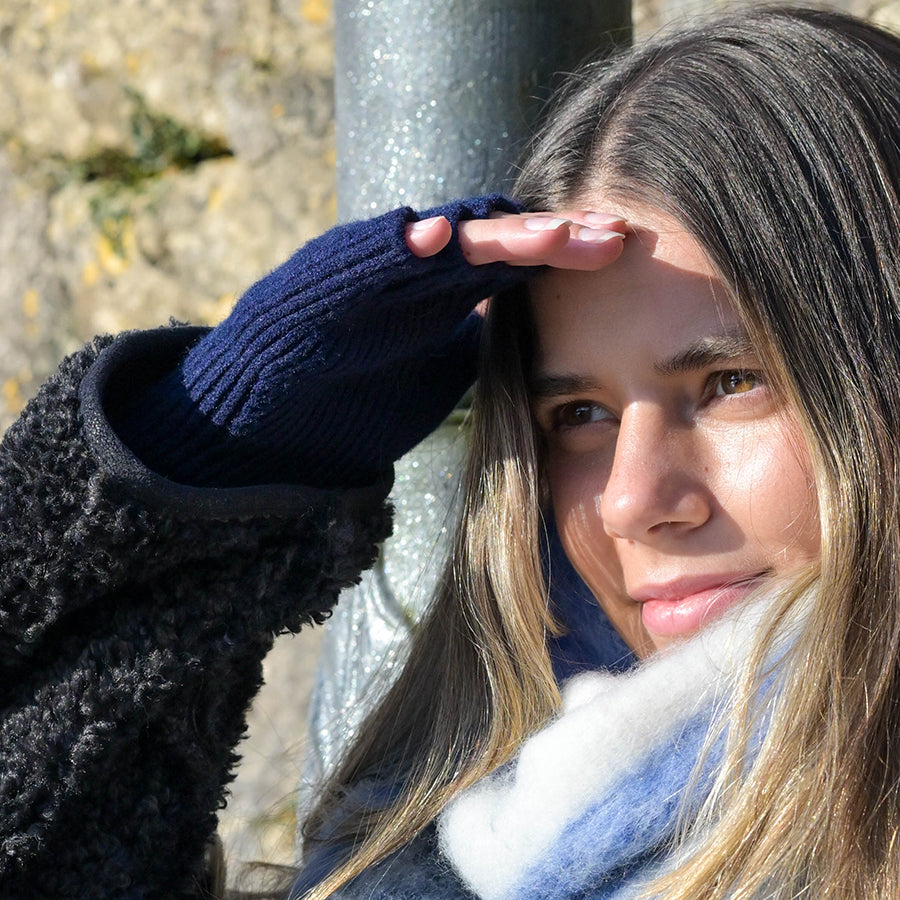 Model shielding eyes from the sun with one hand wearing POM navy fine rib fingerless gloves, styled with a black teddy fleece coat and a pale blue fluffy scarf outdoors against a stone wall.
