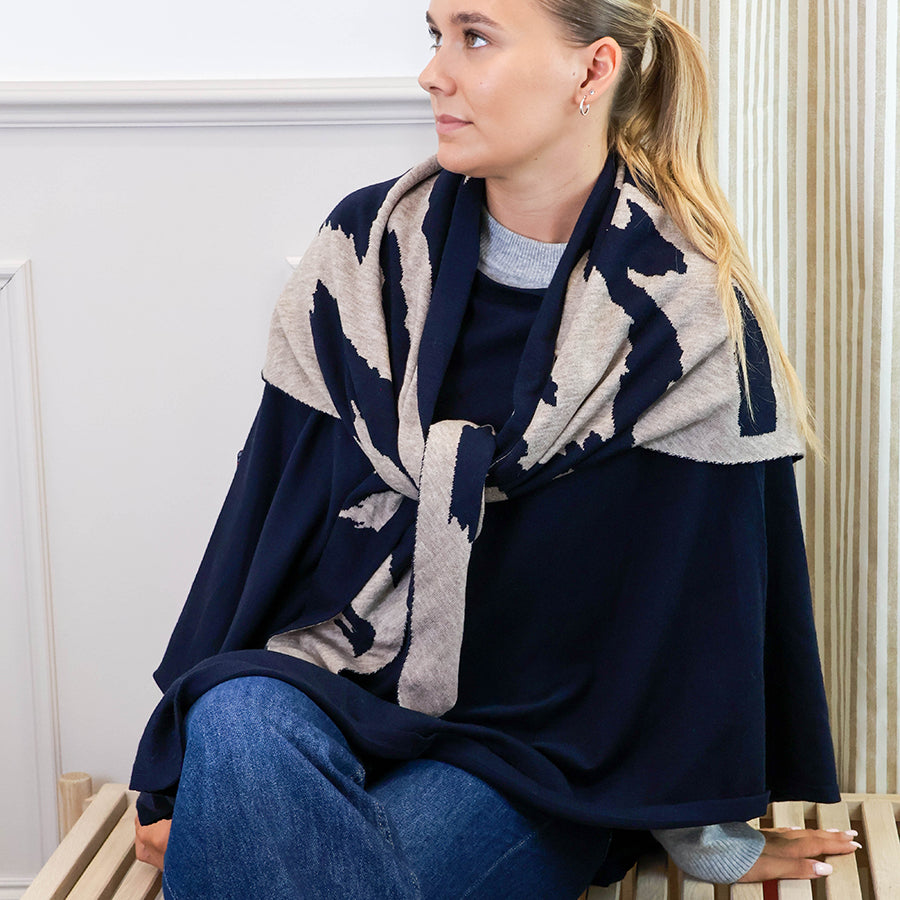 Model seated wearing the POM recycled blend navy poncho with a navy and oatmeal animal print scarf, styled over a grey knit jumper and blue jeans.