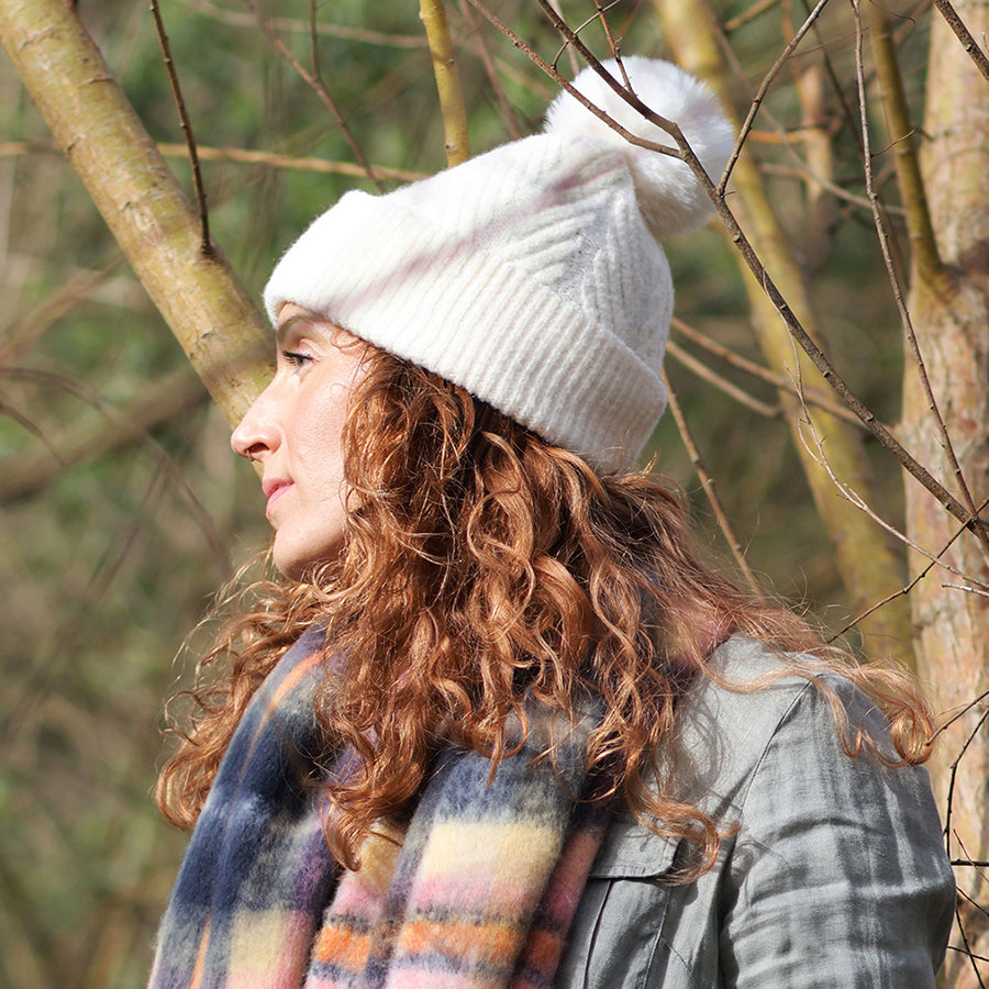 Model wearing POM Charlotte cream bobble hat with faux fur pom pom, styled with a multicolour check scarf and grey jacket outdoors among winter trees.