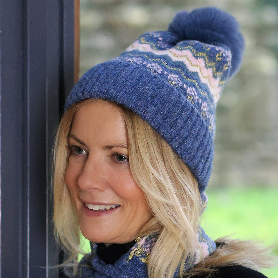 Smiling blonde model leaning against a dark door frame wearing the POM Sofia denim and pink Fair Isle bobble hat with the deep ribbed cuff pulled down, paired with the matching Sofia Fair Isle pull-through scarf, with a green garden behind.