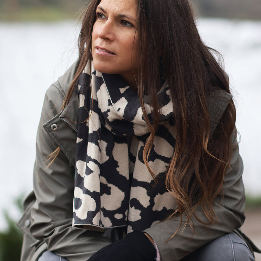 Model seated outdoors by a lake looking to the side wearing the POM Carly oatmeal and black animal print knitted scarf wrapped around her neck, styled with a khaki waxed jacket and dark jeans.