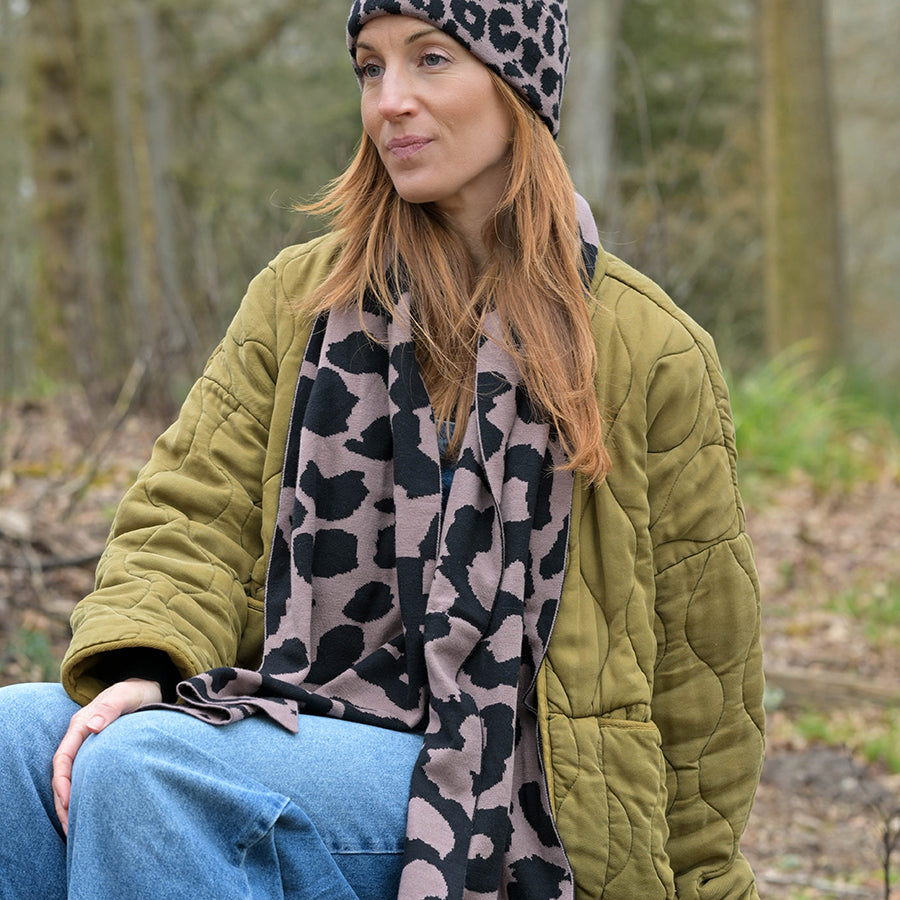 Model sitting outdoors wearing the POM Carly taupe and black animal print scarf with a matching animal print beanie hat, styled over a khaki quilted jacket and wide-leg jeans.