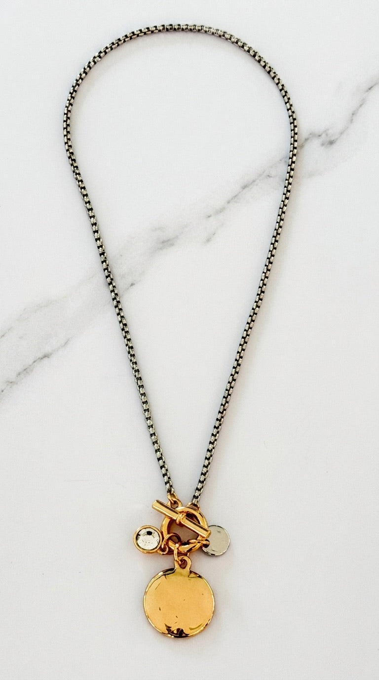 Envy dark hematite box chain necklace with gold T-bar toggle clasp and triple charm cluster of hammered gold coin, crystal and silver disc on marble background