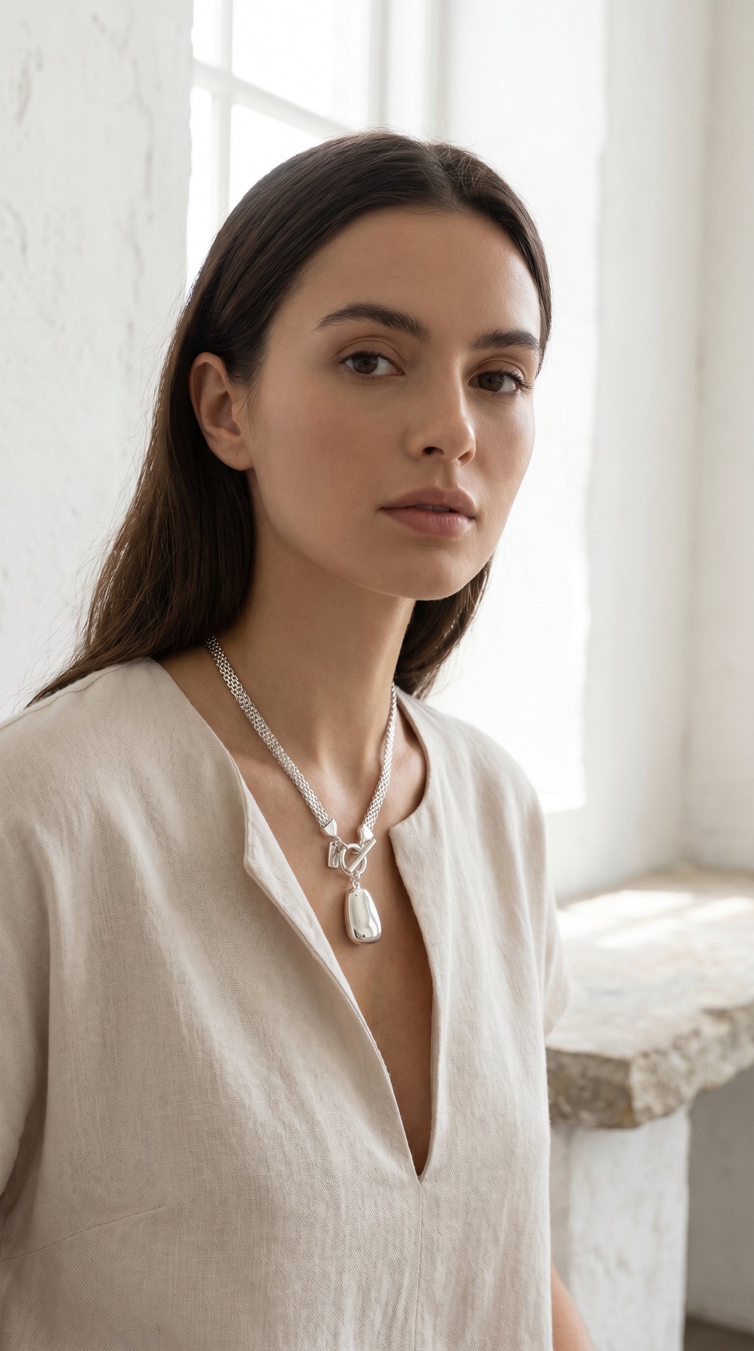 Model wearing Envy silver double mesh chain T-bar necklace with pebble pendant with cream linen v-neck top