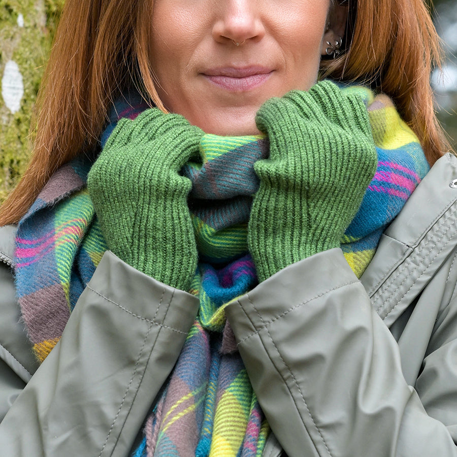 Model wearing POM Yasmin grass green ribbed fingerless gloves, hands raised to chin, styled with a vibrant green, blue, pink and yellow tartan scarf and a khaki jacket outdoors.