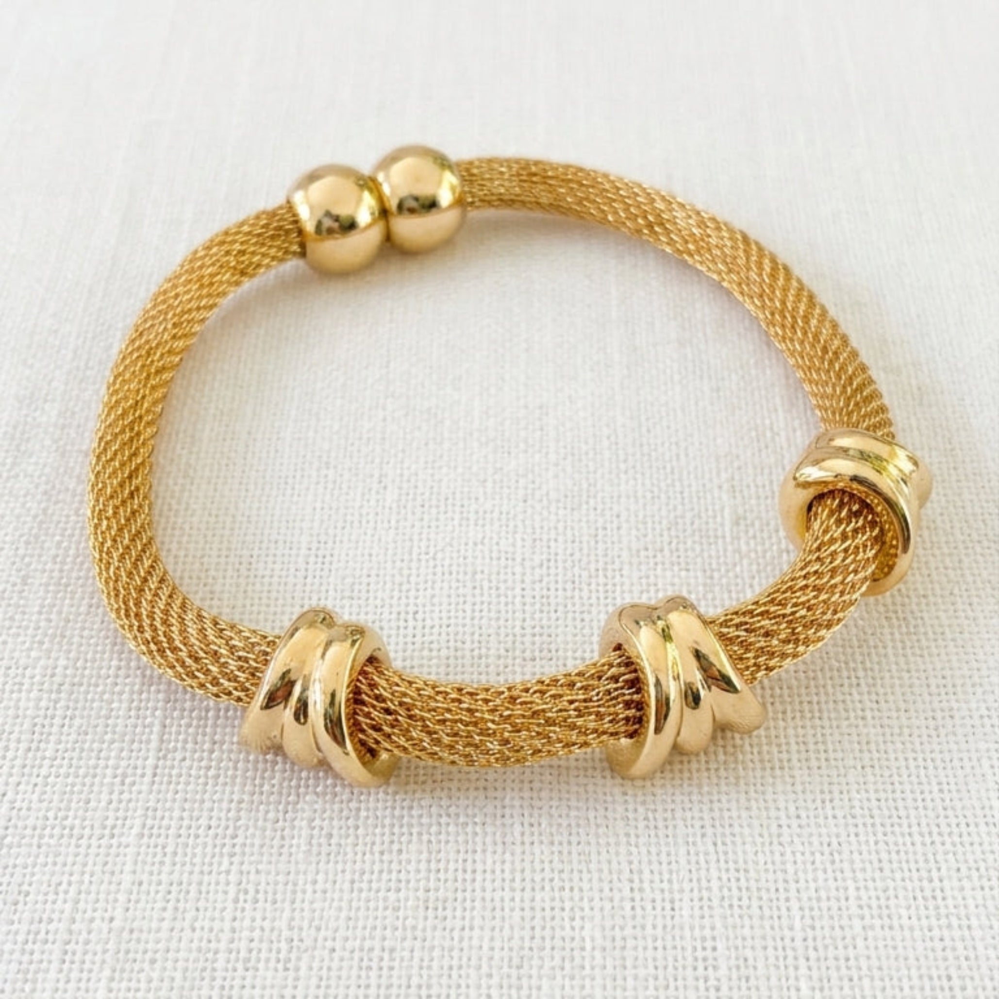 Envy gold-colored mesh bracelet with polished gold bead accents and three knotted wrap details, displayed on a textured off-white fabric background.