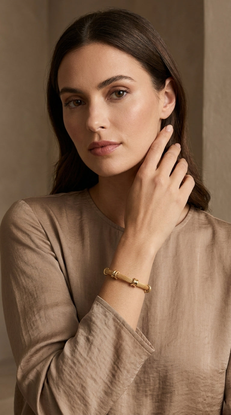 A woman wearing a gold-toned beaded bracelet with a central textured spacer, styled with a taupe linen top against a neutral background.