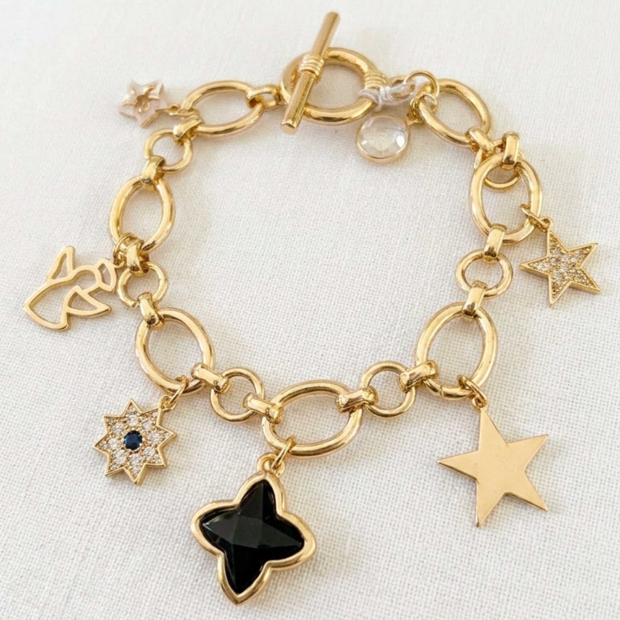 Envy gold charm bracelet featuring star and heart charms, including a prominent black star charm, displayed on a textured white fabric background.