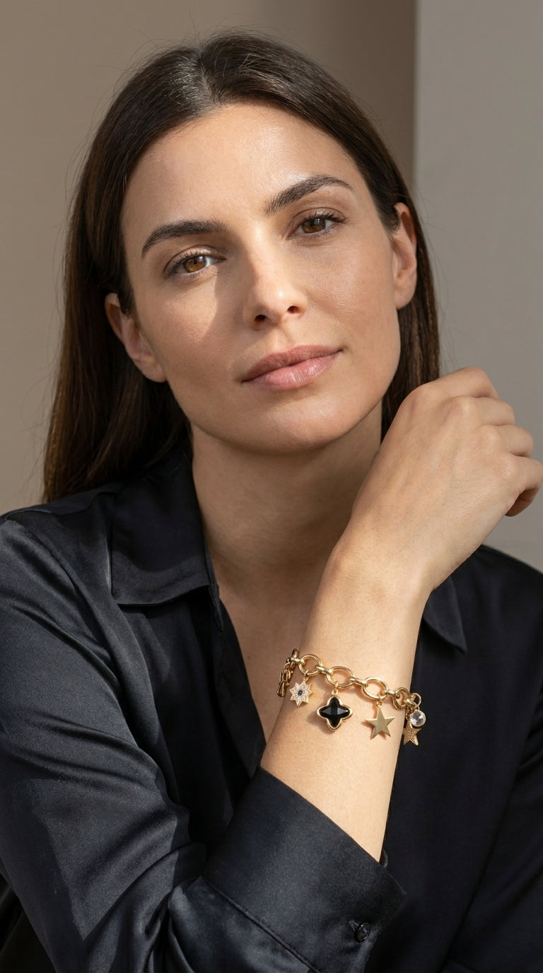 A woman wearing a gold charm bracelet with star and clover charms, styled with a black satin shirt against a neutral beige background.