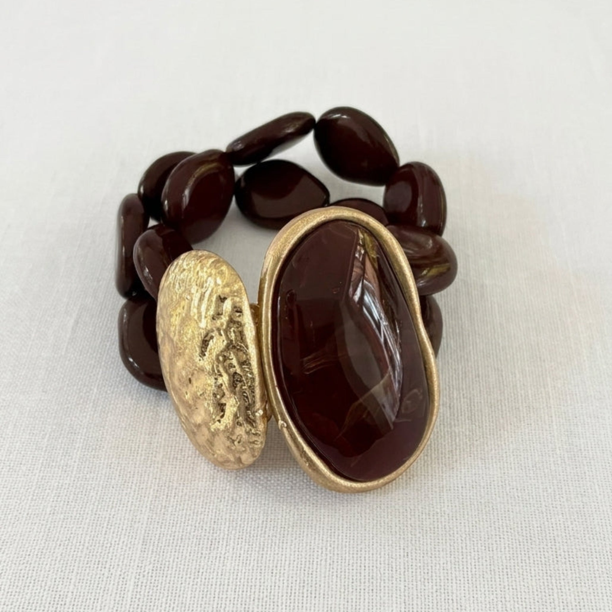 Envy stretch bracelet featuring dark brown polished stone beads, a large oval dark brown cabochon with a gold-tone bezel, and a textured gold-tone oval accent, displayed on a white fabric background.