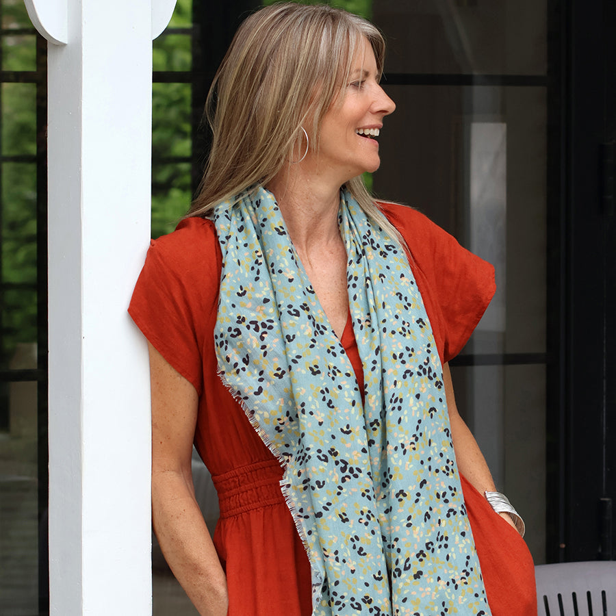 Smiling model leaning against a white pillar outdoors wearing the POM pale mint green ditsy print scarf draped loosely around the neck, styled with a rust red short-sleeve dress and gold hoop earrings.