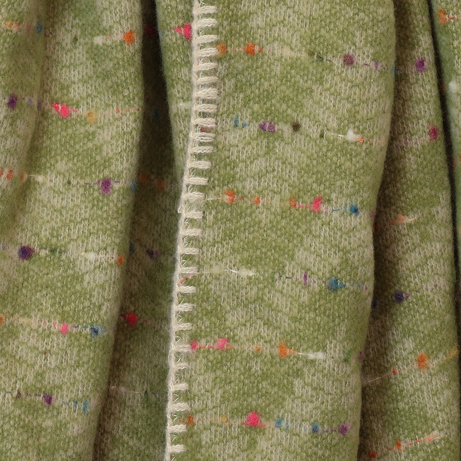 Close-up of the POM Tara pistachio green confetti fleck blanket wrap fabric, showing the scattered pink, orange, blue and purple confetti fleck on the soft pistachio green weave with cream chevron blanket stitch edge detail.