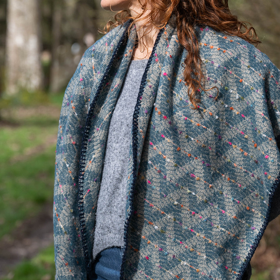 Model with curly auburn hair standing in a sunny woodland clearing wearing the POM Tara teal mix chevron weave blanket wrap draped over the shoulders as a shawl, styled with a grey knit jumper and jeans, with dappled sunlight and trees behind.