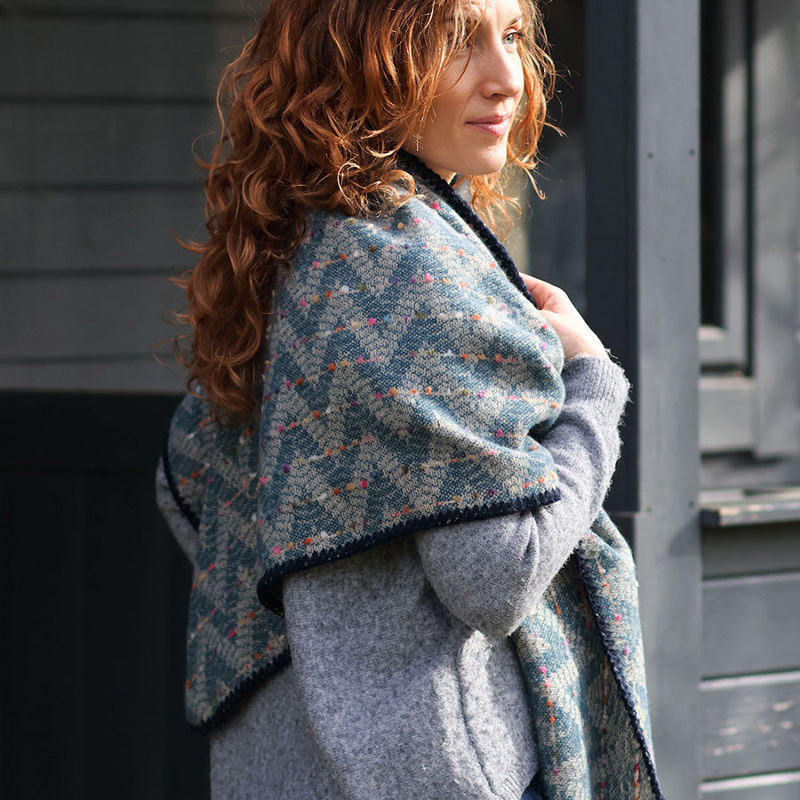 Model with curly auburn hair standing in profile on a sunlit street wearing the POM Tara teal mix chevron weave blanket wrap wrapped around the shoulders, styled with a grey knit jumper, with a dark wooden building behind.