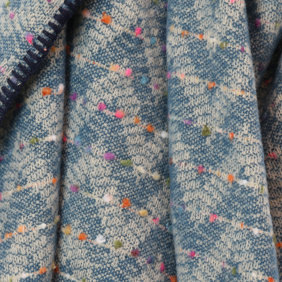 Close-up of the POM Tara teal mix chevron weave blanket wrap fabric, showing the teal and ecru chevron weave with scattered pink, orange, green and purple confetti fleck detail and navy blanket stitch edge at the side.