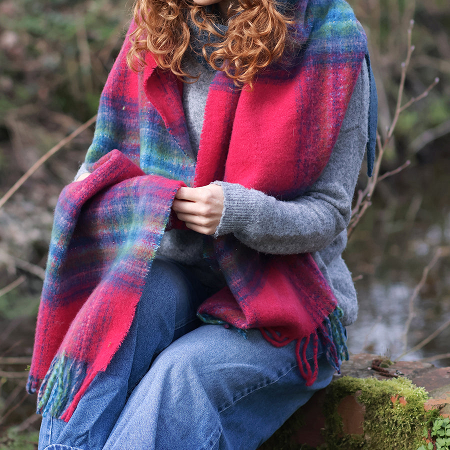 Model with curly auburn hair seated on a mossy stone wall in a woodland setting, wrapped in the POM Miranda blue, green and raspberry mix scarf draped over the shoulders, styled with a grey knit jumper and wide-leg jeans, with a stream and bare winter trees behind.