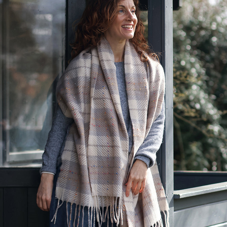 Smiling model with curly auburn hair leaning against a glass door outdoors wearing the POM Olivia taupe mix tartan weave scarf draped loosely over the shoulders as a wrap, styled with a blue-grey knit jumper and jeans, with green trees behind.