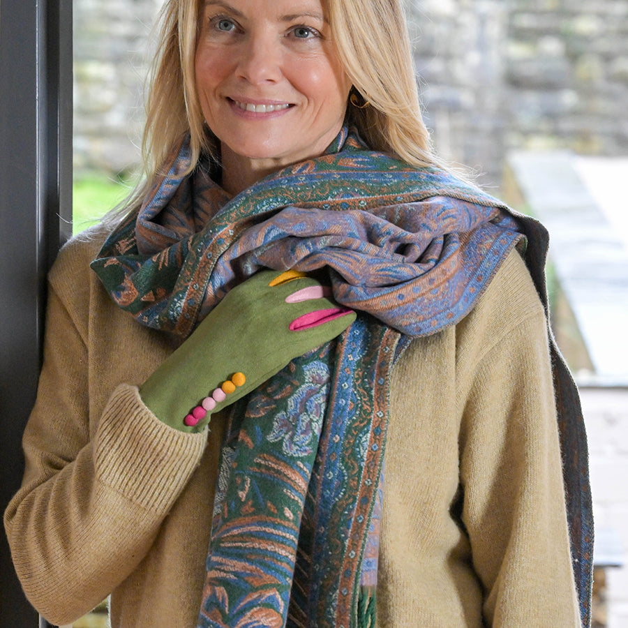 Smiling blonde model leaning in a doorway wearing the POM Celia green-blue and rust reversible jacquard scarf wrapped loosely around the neck showing both sides, styled with a camel knit jumper and olive green fingerless gloves with colourful bobble detail, with a stone wall behind.