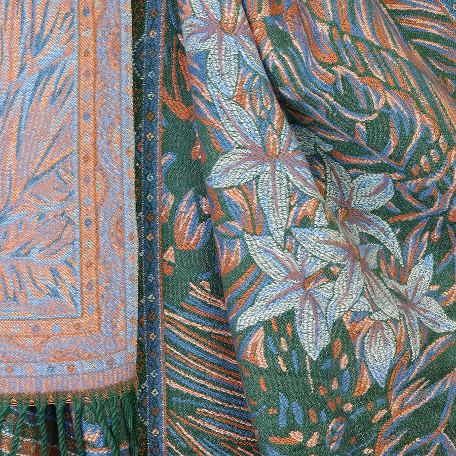 Close-up of the POM Celia reversible jacquard scarf showing both sides: the deep teal green botanical jacquard with large floral and leaf motifs in rust and pale blue on the right, and the soft dusty blue-grey reverse with intricate paisley and geometric border pattern on the left, with twisted green tassel fringe at the bottom.