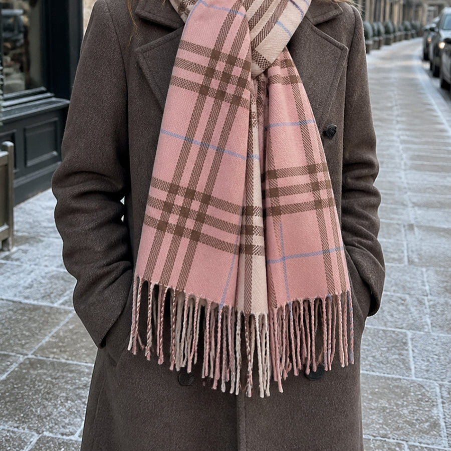 Model standing on a cobbled Parisian street wearing the POM Nellie salmon pink, blue and brown mix check scarf tied loosely at the front, styled with a dark brown tailored wool coat, with a tree-lined boulevard behind.