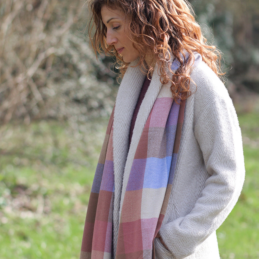 Model looking down outdoors in a sunny garden wearing the POM Esme blush and blue mix check scarf wrapped around the neck, styled with a cream herringbone coat, with bare winter branches in the background.