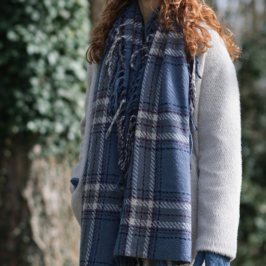 Close-up of model outdoors wearing the POM Darcie dusky blue mix fringe edge check scarf draped over the shoulders and chest, showing the dusky blue, cream and burgundy check pattern with long twisted fringe detail, styled with a cream herringbone coat and ivy-covered wall behind.