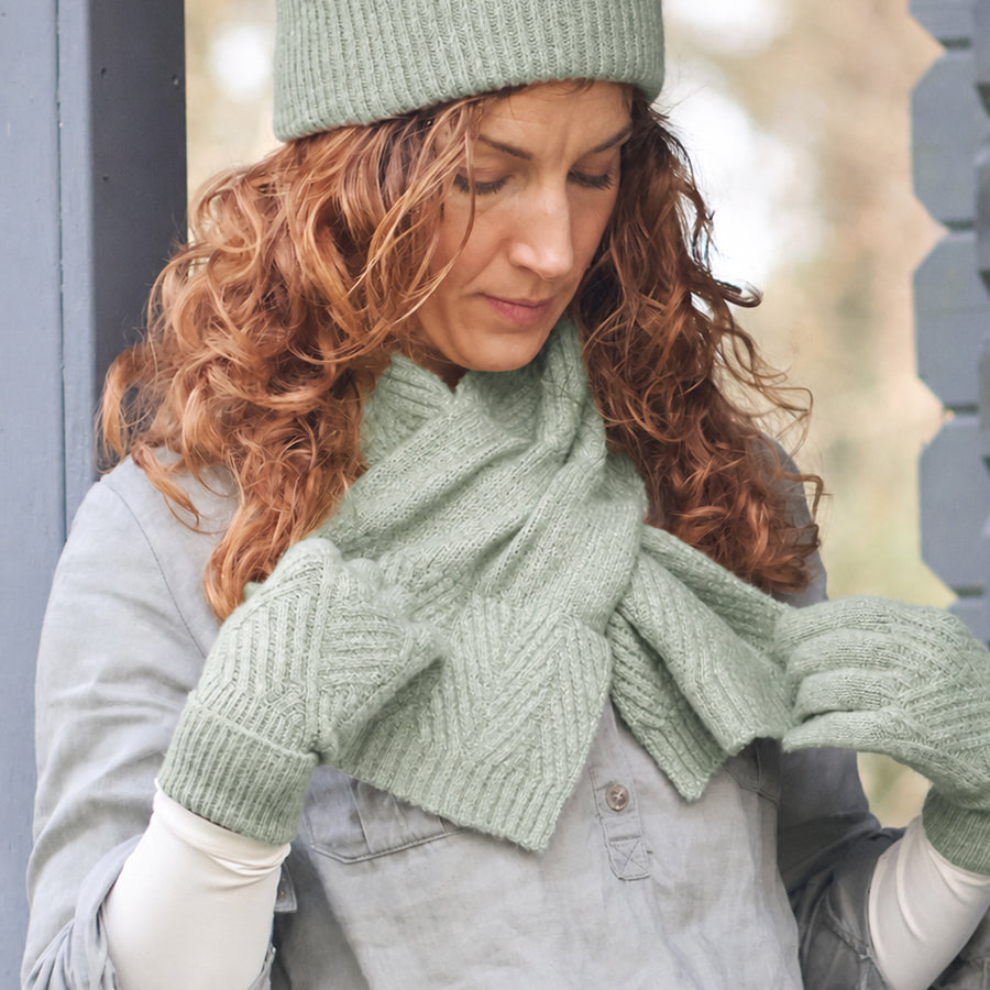 A woman wearing a light sage green knit beanie, matching knit scarf, and knit mittens, standing outdoors near a gray wooden structure.