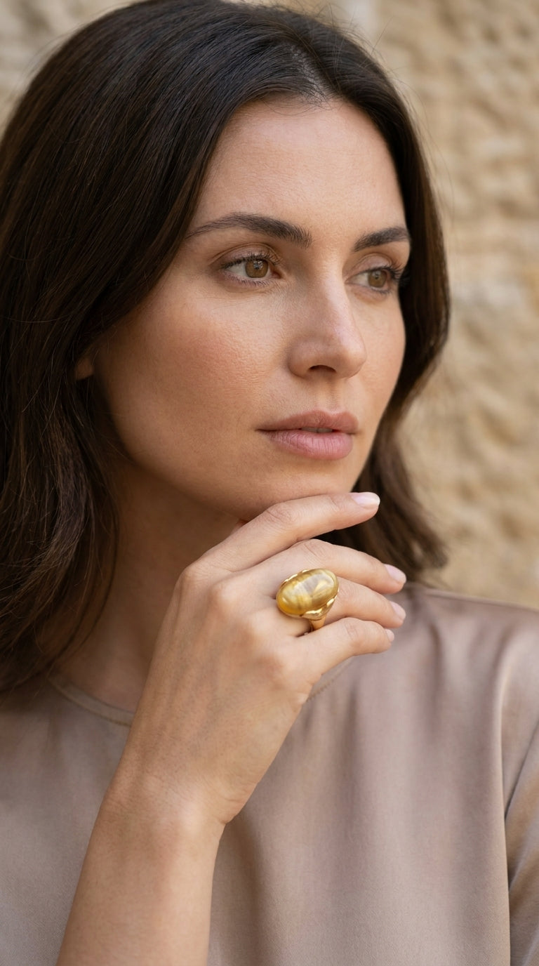 A close-up lifestyle shot of a woman with dark hair wearing a large, chunky gold ring with a round white stone, set against a soft beige background.